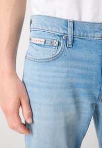 Svijetloplave traperice s srednjim strukom, šivanjem, i crvenom oznakom Calvin Klein na stražnjem džepu.