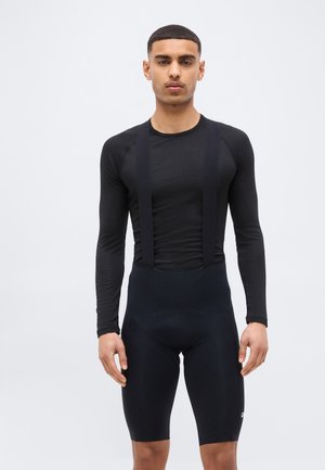 Mladi moški, oblečen v črno dolgoročno športno majico in črne kolesarske hlače z naramnicami, stoji pred enostavnim svetlim ozadjem.