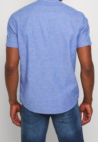 Hombre con una camisa de manga corta azul clara con pequeños puntos azul oscuro y jeans azules, visto desde atrás contra una pared lisa.