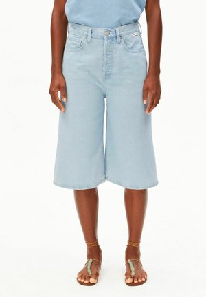 Personne portant un short en denim bleu clair à jambes larges et des sandales à lanières avec des détails en perles vertes, debout devant un fond blanc.