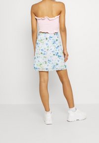 Rosa gesmoktes Trägertop mit rüschigem Saum, kombiniert mit einem blauen Minirock mit floralen Mustern. Das Model trägt weiße Sneakers und ist mit dem Rücken zur Kamera gewandt.