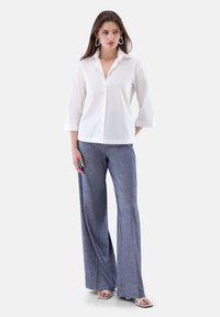 White button-up shirt with a collar and three-quarter sleeves, paired with wide-leg blue patterned trousers. Model also wears silver earrings.