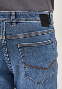 Blaue Jeans aus Denim mit tailliertem Schnitt, ausgestattet mit orangefarbenen Nähten, zwei Gesäßtaschen und einem schwarzen, markierten Lederpatch an der Taille.