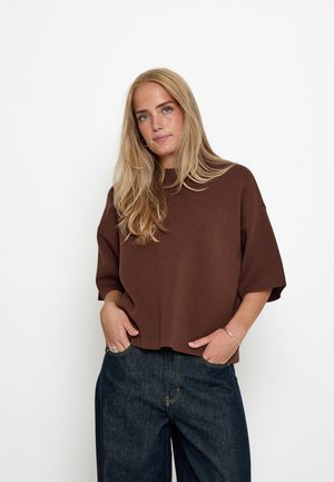 Junge Frau mit langen blonden Haaren trägt ein lockeres braunes Oberteil und dunkelblaue Jeans, steht mit den Händen in den Taschen vor weißem Hintergrund.