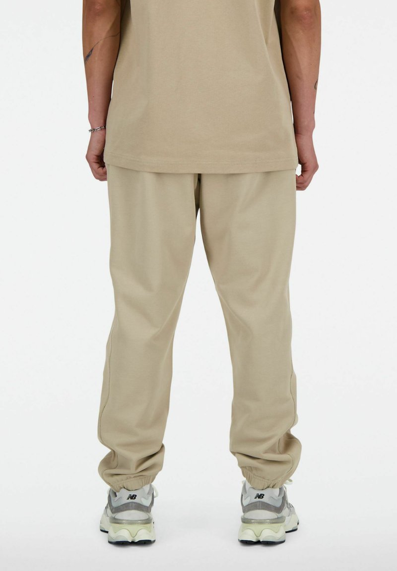 Joggers en coton beige avec des poignets élastiques et une coupe décontractée, assortis à des baskets grises et blanches. Présente un design simple sans motifs.