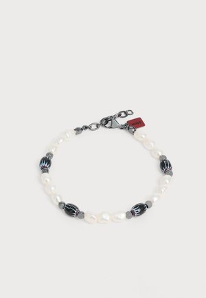 Armbånd med hvide uregelmæssige perler, sorte ovale perler med blå og røde mønstre, grå afstandskugler, en karabinlås og et rødt Hugo-mærke.