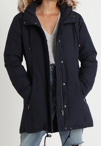 Mörkblå, isolerad parkas med huva som har pälsdetaljer. Har sidofickor, dragkedja framtill och justerbar dragsko i midjan.