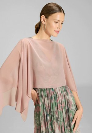 Frau trägt einen transparenten rosa Umhang über einem geblümten, plissierten Kleid, posiert mit zurückgebundenem Haar und Creolen vor einem schlichten Hintergrund.