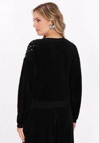 Zwarte fluwelen sweatshirt met lange mouwen, ribbelmanchetten en versierde schouderdetails met metalen accenten. Soepele textuur en een losse pasvorm.