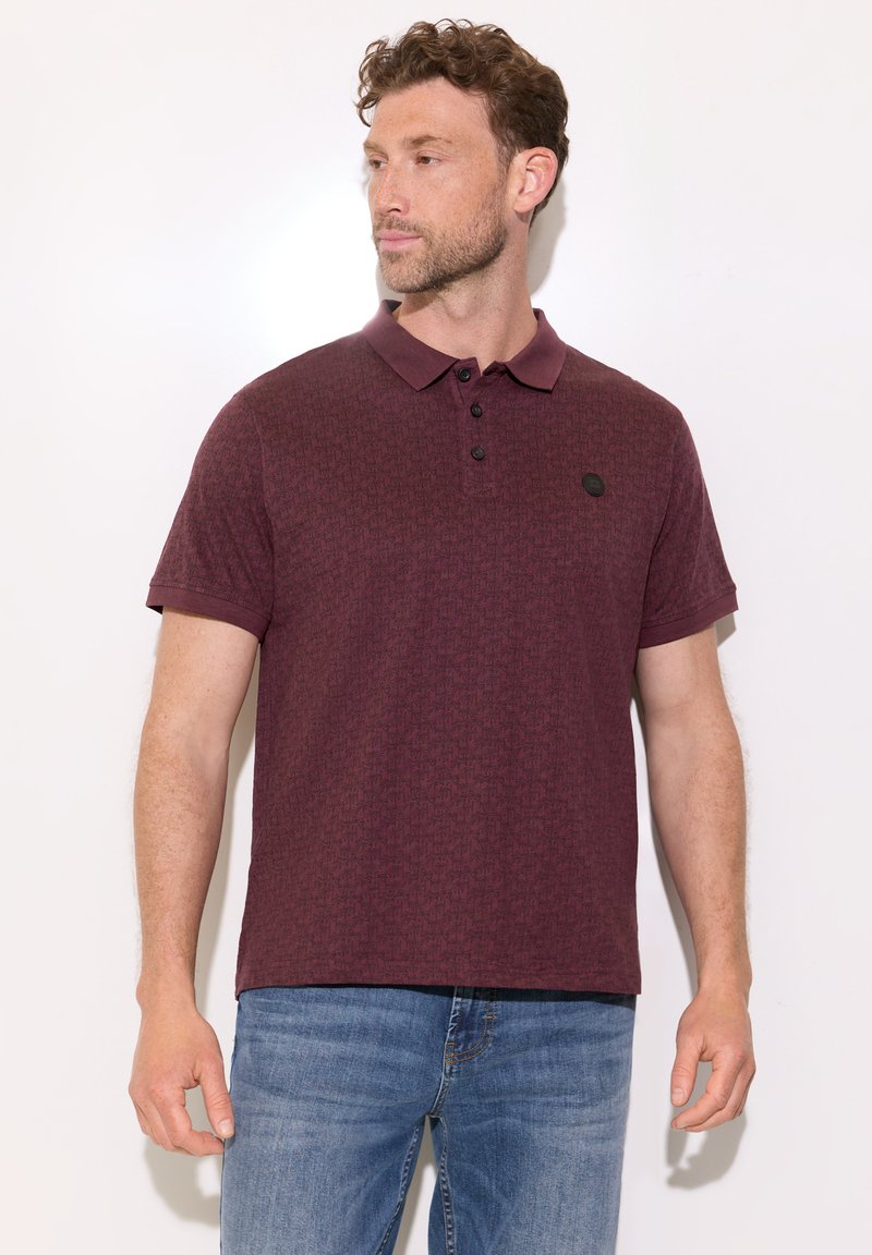 Homme portant un polo rouge foncé à manches courtes et un jean bleu, regardant sur le côté sur un fond blanc uni.