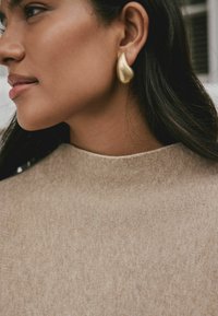 Pendiente de lágrima dorado en la oreja de una mujer, acentuado por un suéter beige de cuello alto con una textura suave.