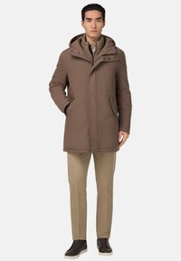 Manteau en mélange de laine marron avec capuche, doté d'un zip sur le devant, de fermetures à pression et de poches latérales. Porté sur un gilet beige et un pantalon de couleur claire.