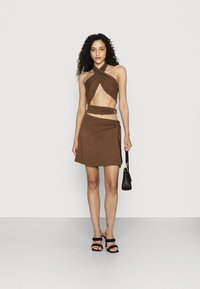 Conjunto de blusa cruzada em tom castanho e saia mini em tecido suave, com alças cruzadas e detalhe de nó lateral, acompanhado de uma pequena mala preta.