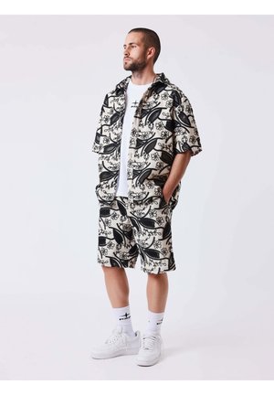 Hombre de pie con las manos en los bolsillos, vistiendo una camisa de manga corta y pantalones cortos a juego con estampado floral en blanco y negro, zapatillas blancas y calcetines blancos.