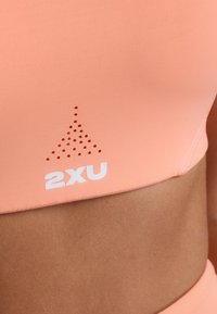 Perzik sportbeha met een gladde textuur, voorzien van perforatie in een driehoekig patroon en een wit "2XU" logo aan de onderlinkerzijde.