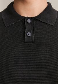 Zbliżenie na kołnierz czarnej dzianinowej koszulki polo z dwoma guzikami i widoczną teksturą materiału na szyi i górnej części klatki piersiowej osoby.