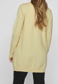 Lång, ljusgul stickad kofta med ribbade ärmslut och nederkant. Mjuk textur, avslappnad passform och inga synliga mönster eller hårdvarudetaljer.