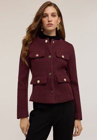 Burgundy tekstuuri jakk krae, neli esist pocketit ja kuldse tooniga nupud, koos musta särgi ja täpsete mustade pükstega.