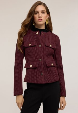 Veste texturée bordeaux avec un col, quatre poches avant et des boutons dorés, associée à un haut noir et un pantalon noir ajusté.