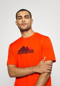 Červené tričko s krátkým rukávem značky Helly Hansen, na přední straně s černým logem a grafikou hor. Hladký materiál se kulatým výstřihem.