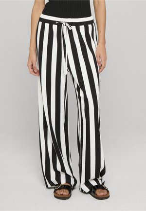 Kobieta ubrana w czarno-białe szerokie spodnie w pionowe paski, ze sznurkiem w pasie i czarne sandały.