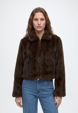 Femme aux longs cheveux ondulés, vêtue d'une veste en fourrure marron foncé et d'un jean bleu, debout devant un fond clair uni.