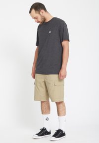 Camiseta gris de manga corta con un pequeño logo, combinada con pantalones cortos tipo cargo beige con bolsillos laterales, zapatillas negras y calcetines blancos con diseño.
