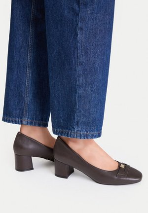 Benen met donkerblauwe spijkerbroek en bruine schoenen met een lage blokhak en een klein decoratief metalen detail bovenop.