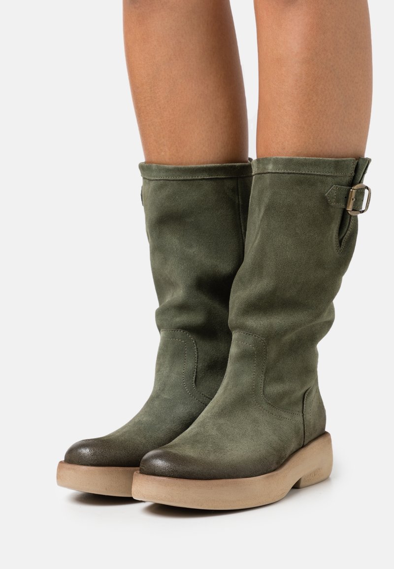 Bottes en suède vertes jusqu'au genou avec un bout arrondi, un capuchon texturé de couleur foncée, et une boucle latérale pour ajustement. Semelle épaisse beige.