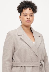 Cappotto beige in misto lana con colletto a scialle, vita con cintura e tasche anteriori. Tessuto morbido e vestibilità su misura, con dettagli di design minimalisti.