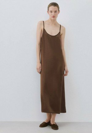 Kvinde stående mod en hvid væg iført en enkel brun ærmeløs maxikjole og matchende brune slip-on sko.