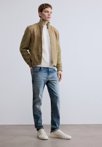 Tégla színű szarvasbőr bomber dzseki bordázott ujjakkal, fehér bordázott pulóver, világoskék, kopott farmernadrág és krémszínű sportcipő. Egyszínű háttér.