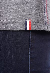 Šedá bavlněná látka má jemnou strukturu, s viditelným červeným, bílým a modrým pruhovaným akcentem na lemu proti tmavě modrému denimu.