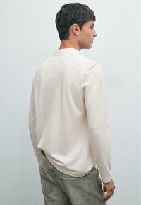 Langärmliger, cremefarbener Pullover aus weichem Material mit klassischem Kragen und einem einfachen, geradlinigen Design. Verwaschene graue Jeans ergänzen das Outfit.