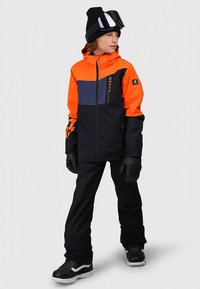 Skidjacka i orange och marinblå färgblock, tillverkad av vattentätt material; kombinerad med svarta byxor och handskar; snowboardglasögon på huvudet.