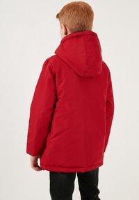 Punainen toppatakki hupulla, jossa on sileä pinta ja suora leikkaus, nähtynä takaa. Mustat housut näkyvät alapuolella.