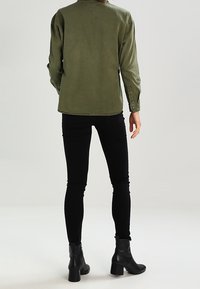 Camisa de manga larga verde oliva con un cuello sencillo, llevada sobre pantalones negros ajustados y botas negras hasta el tobillo, vista desde la parte de atrás.