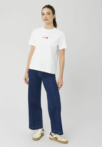 T-shirt en coton blanc à manches courtes avec un logo graphique rouge sur la poitrine, associé à un jean bleu foncé ample et des baskets claires.