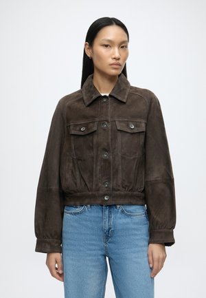Mujer con el cabello liso y negro, que lleva una chaqueta de ante marrón oscuro con botones y vaqueros azul claro, de pie contra un fondo claro y sencillo.