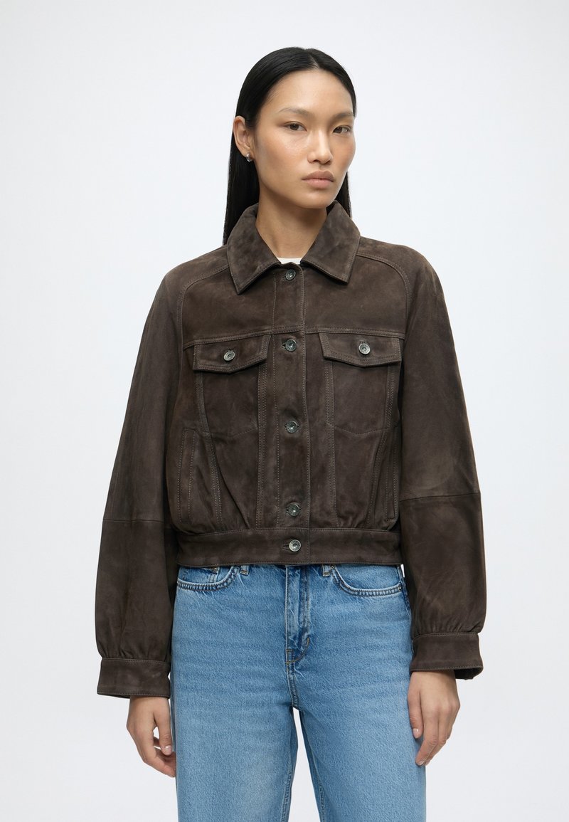 Femme aux cheveux noirs lisses portant une veste boutonnée en daim marron foncé et un jean bleu clair, debout devant un fond clair uni.