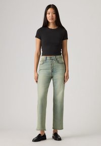 Μαύρο τοπ με κοντά μανίκια, ανοιχτοπράσινο τζιν ψηλόμεσο, ίσιο παντελόνι, ξεθωριασμένο φινίρισμα, συνδυασμένο με μαύρα μοκασίνια, μίνιμαλ αξεσουάρ, ουδέτερο φόντο.