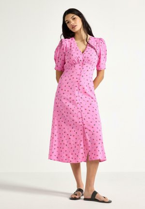 Kvinde iført en lyserød, knappet midikjole med pufærmer og lille blomsterprint, stående med hænderne bag ryggen og sorte slip-in sandaler.