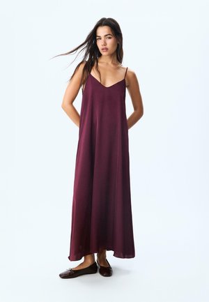 Robe maxi bordeaux avec un décolleté en V, des fines bretelles et une coupe fluide. Tissu lisse et léger qui tombe jusqu'aux chevilles. Assortie à des ballerines noires.