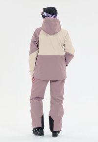 Skidjacka och byxset i ljuslila och beige, med en huva, justerbara ärmslut och svarta detaljer vid byxans fållar.