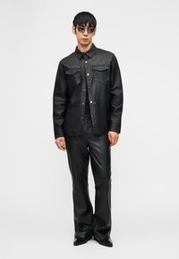 Άνδρας που φοράει μαύρο δερμάτινο πουκάμισο με κουμπιά, ταιριαστά δερμάτινα παντελόνια, μαύρα παπούτσια και σκουρόχρωμα γυαλιά ηλίου, στέκεται μπροστά σε μονόχρωμο φόντο.