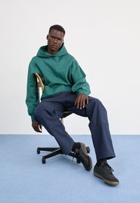 Zielona bluza z kapturem, luźne granatowe spodnie, czarne sneakersy z gumową podeszwą. Temat siedzący na obrotowym krześle na jednolitym tle.