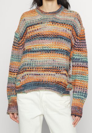 Personne portant un pull en maille rayé multicolore avec des tons orange, bleu, vert et rose, associé à un pantalon blanc.