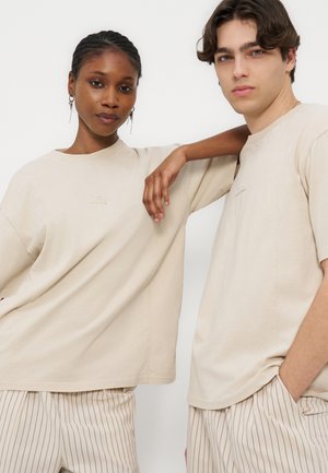 Δύο νέοι ενήλικες φοράνε μπεζ μπλούζες και ριγέ παντελόνια, ο ένας με το χέρι του ακουμπισμένο στον ώμο του άλλου, κοιτάζοντας μπροστά με φόντο ένα λευκό φόντο.