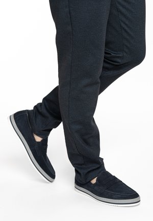 Lower legs in dark slim-fit trousers and dark slip-on shoes with white soles, one foot slightly crossed over the other.