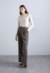 Beige lange mouw top gecombineerd met hoge taille grijze geplooide broek en bruine sneakers met witte zolen, tegen een effen achtergrond.